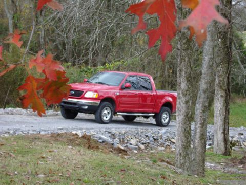 2007 Ford Mustang - A red pickup truck is seen from a 3/4 front angle on a gravel road, surrounded by trees with fall foliage.