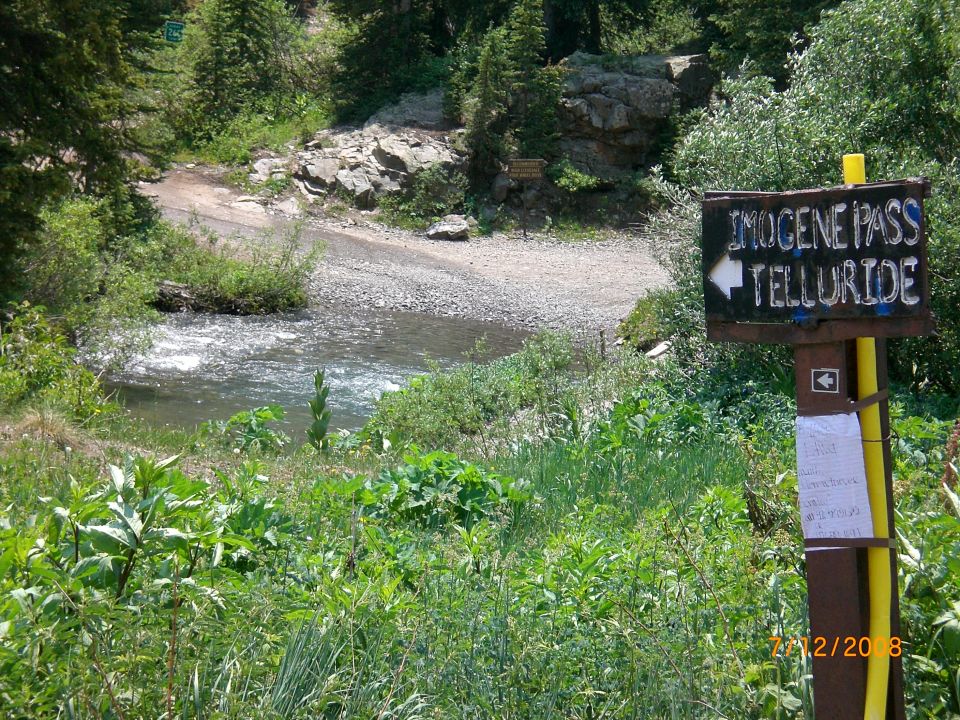 Sign pointing to Imogene Pass and Telluride next to a river in a mountainous area