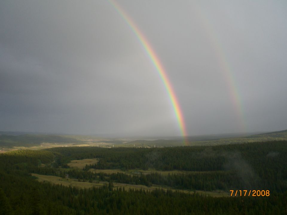Double rainbow arcs across a cloudy sky over a vast forest and valley