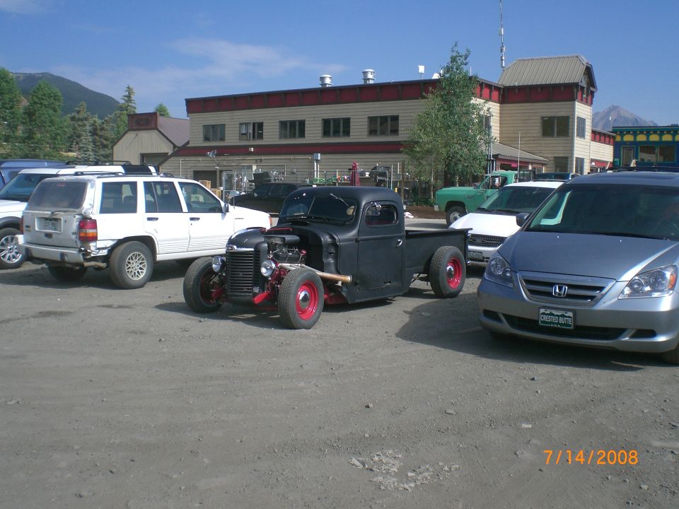 Black hot rod pickup truck with exposed engine and red wheels, parked in a lot
