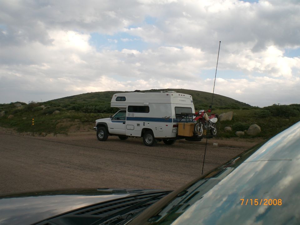 Truck camper parked on dirt road with a motorcycle on the back. Hill in background