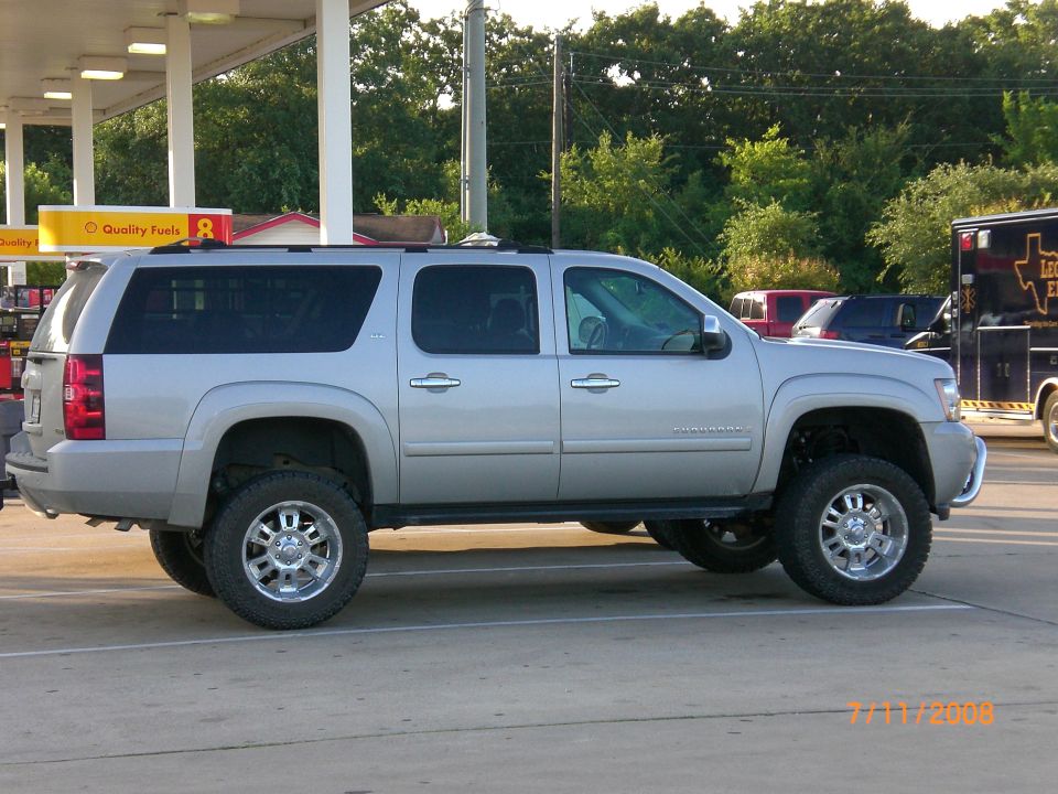 Silver SUV, looks like a Suburban, with big tires and lifted suspension. Parked at a gas station