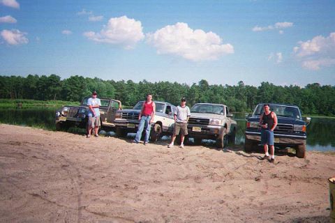1990 Ford Bronco - Four people stand in front of four off-road vehicles parked on a sandy area near a body of water and a treeline.