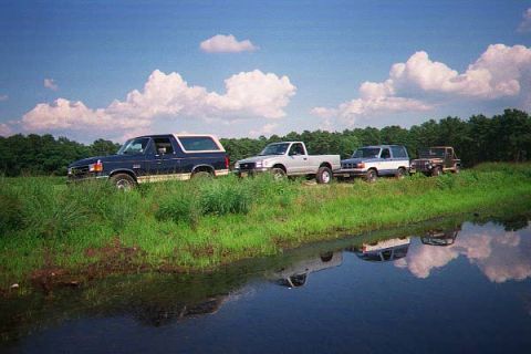 1990 Ford Bronco - A line of four SUVs and trucks are parked on a grassy embankment next to a body of water. The vehicles are a dark blue SUV, a silver pickup…