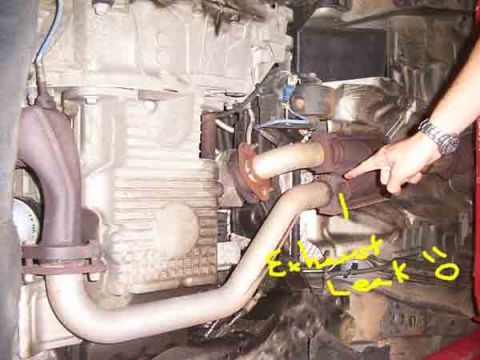 1998 Ford Taurus - A hand points to an exhaust leak on the underside of a vehicle, with yellow marker highlighting the area.