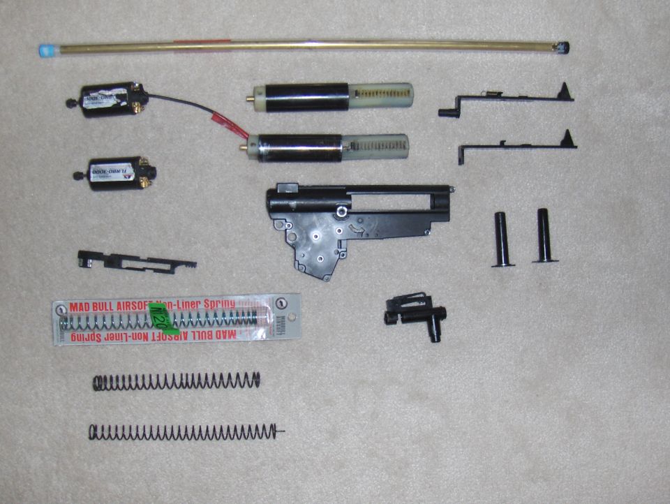 Airsoft gun internal parts laid out, including motors, springs, gearbox components, and a barrel