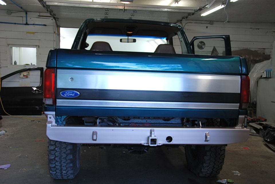 Rear view of a teal Ford Bronco with a silver stripe and a tow hitch. Doors are open