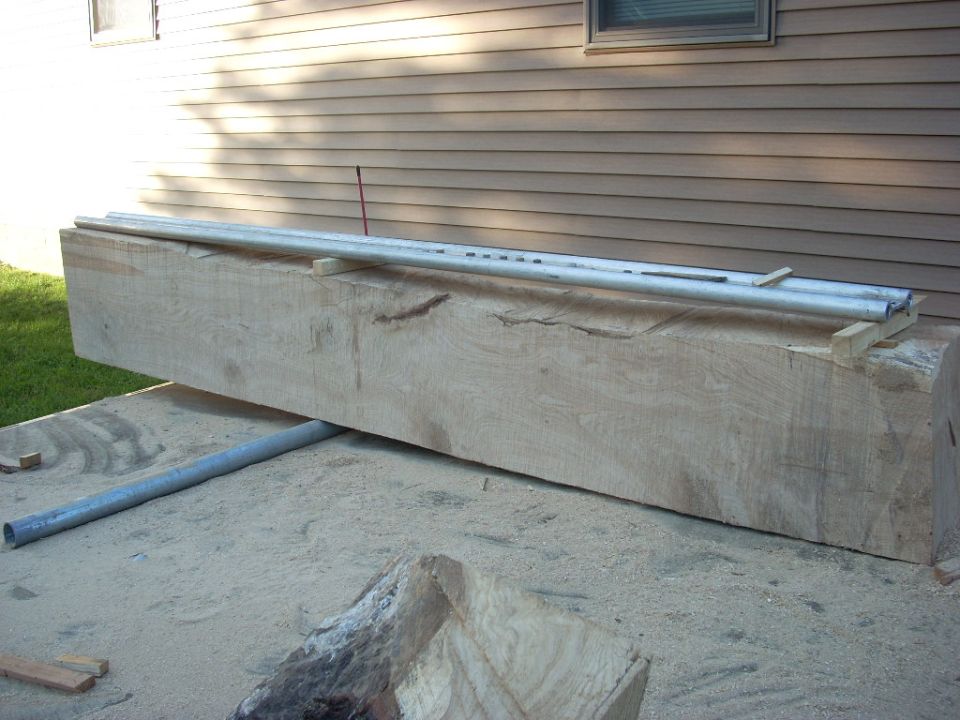 Large wooden beam with metal pipes on top, resting on sand and wood scraps
