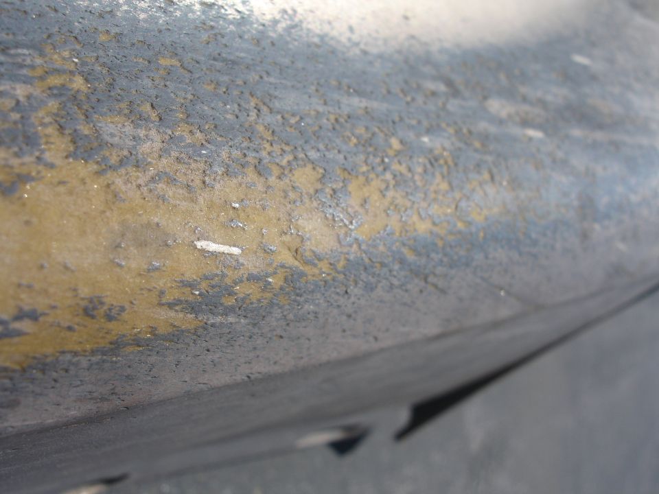 Close-up of weathered, peeling paint on a dark gray car part with yellow undercoat visible