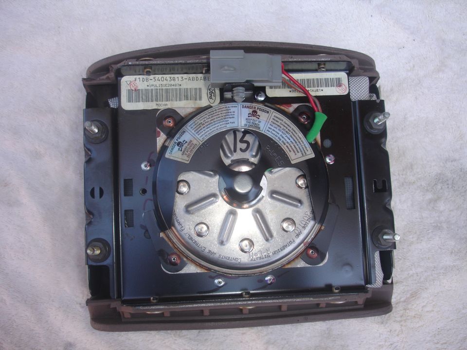 Airbag module with labels and wiring, showing a metal circular component with bolts