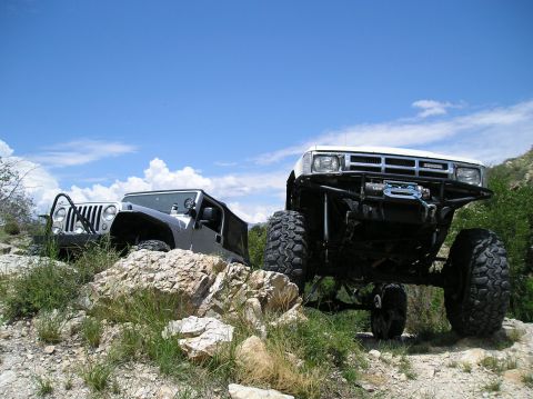 1986 Toyota Hilux - Two modified off-road vehicles navigate a challenging rocky trail under a blue sky.