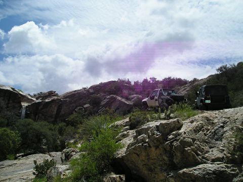 1986 Toyota Hilux - Two off-road vehicles navigate a , rocky terrain with sparse vegetation and a distant waterfall.