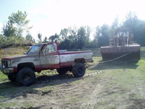 1995 Mercury Grand Marquis - A lifted pickup truck tows a large excavator bucket across a muddy field under a bright sky.