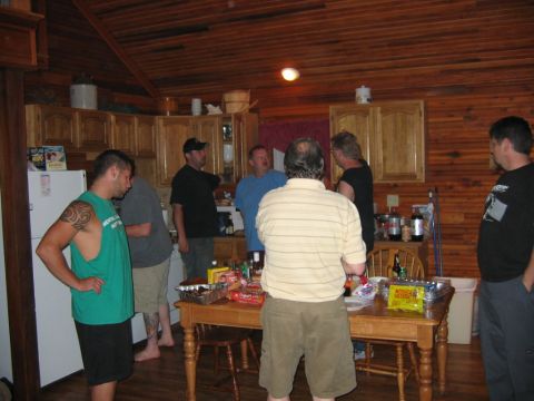 1985 Ford LTD Crown Victoria - A group of men gather in a wood-paneled kitchen, with a table full of snacks and beverages.