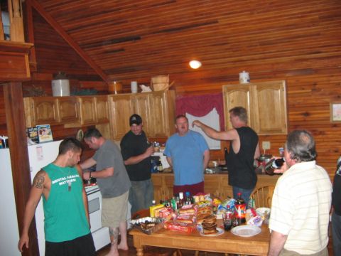 1985 Ford LTD Crown Victoria - A group of men are gathered in a wood-paneled kitchen, with a table full of food and drinks.