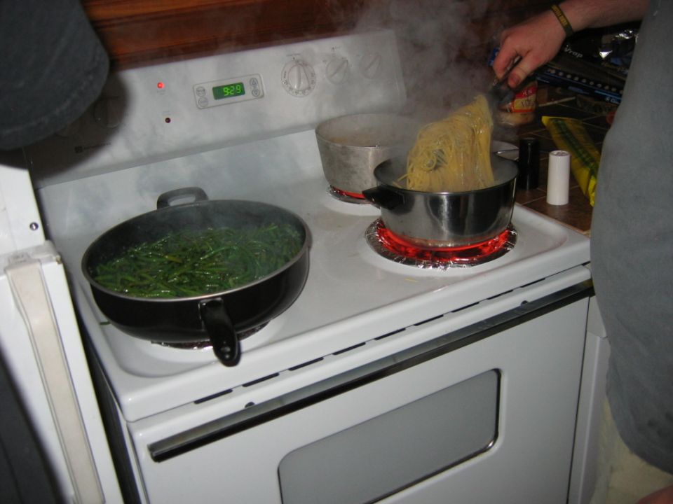 Someone is cooking pasta and green beans on a white electric stove. The pasta is being lifted out of a pot of boiling water