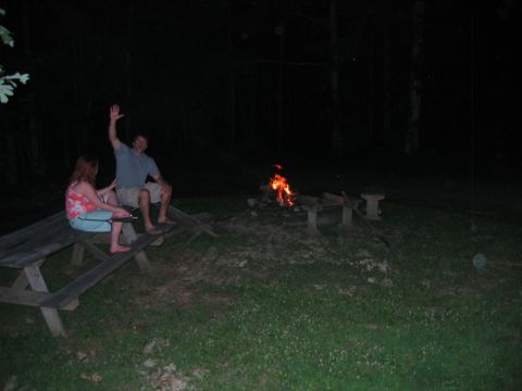 1985 Ford LTD Crown Victoria - A man waves while sitting on a picnic table with a child next to a campfire in the dark.