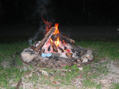 1985 Ford LTD Crown Victoria - A campfire burns brightly at night, casting a warm glow on the surrounding grass and dirt.