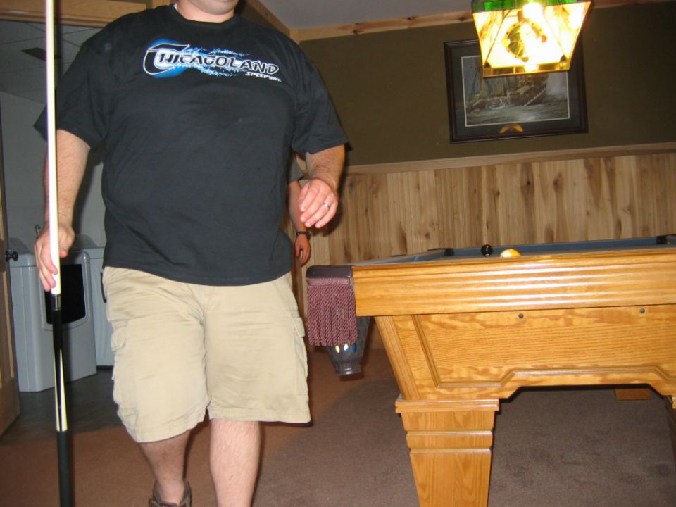 Man in a black t-shirt and khaki shorts holding a pool cue near a pool table