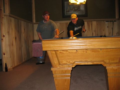 1985 Ford LTD Crown Victoria - Two men are playing a game of pool in a room with wood paneling and a framed picture above the table.