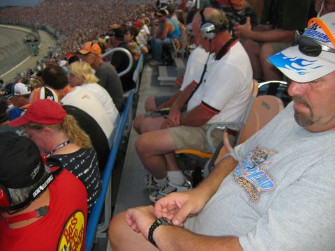 1985 Ford LTD Crown Victoria - A crowd of people watch a race from the stands, with some individuals in the foreground wearing hats and listening with headphones.