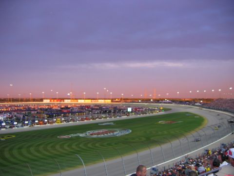 1985 Ford LTD Crown Victoria - A wide view of a NASCAR track and its surrounding parking lot as the sun sets, with lights beginning to illuminate the scene.