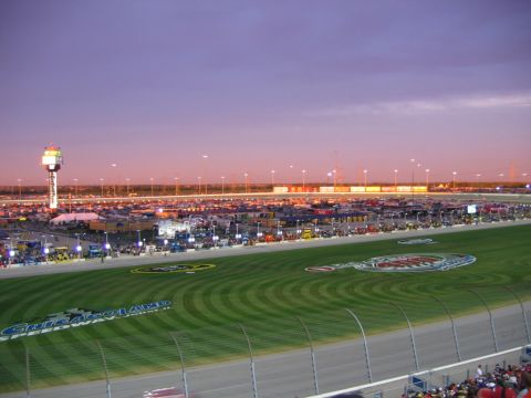 1985 Ford LTD Crown Victoria - A vast expanse of vehicles fills the parking lot surrounding Chicagoland Speedway under a twilight sky.