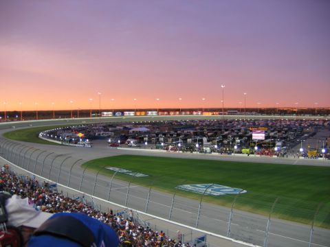 1985 Ford LTD Crown Victoria - A wide view of a NASCAR oval speedway at dusk, showing packed grandstands in the foreground, the racing surface, infield with vehicles and…