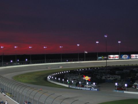 1985 Ford LTD Crown Victoria - Dusk view across the banking at Chicagoland Speedway, with two stock cars running the backstretch under the lights against a deep red sunse…