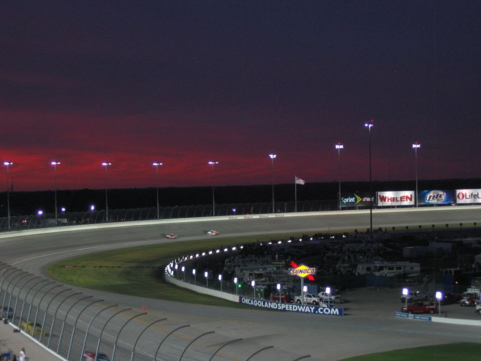 Two stock cars racing at dusk at Chicagoland Speedway under red sunset sky