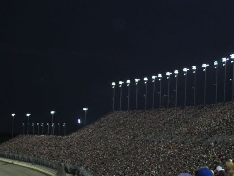 1985 Ford LTD Crown Victoria - A large crowd fills the grandstands of a race track illuminated by numerous tall light poles against the night sky.