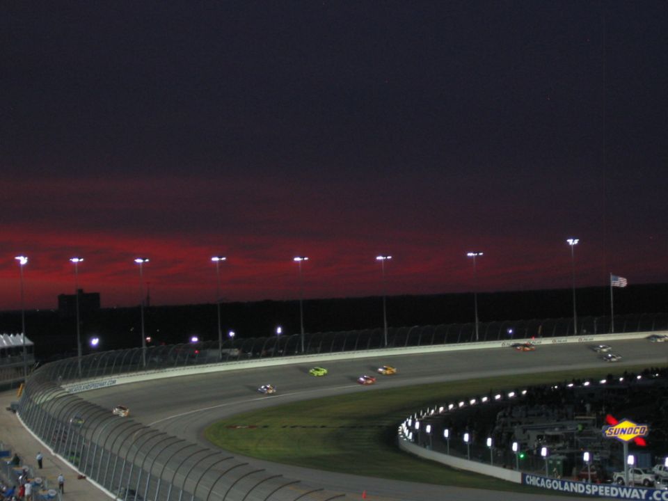 Race cars on a track at dusk with a red sky and stadium lights