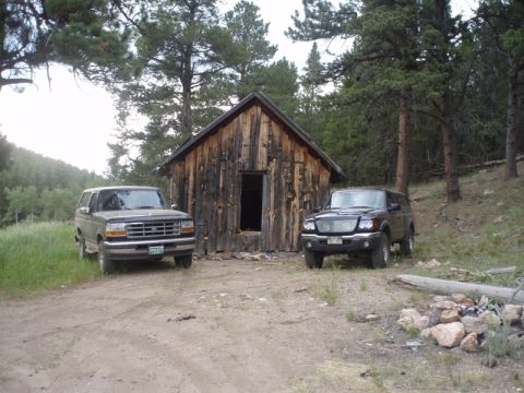 1996 Ford Bronco - Two SUVs are parked outside a rustic wooden cabin in a wooded area.