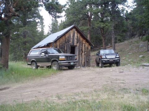 1996 Ford Bronco - A 1996 Ford Bronco and a black Ford pickup truck are parked in a clearing next to an old wooden cabin.