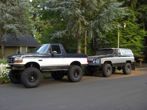 1993 Ford Bronco - Two modified trucks, a Ford and a Chevrolet, are parked on a paved surface in front of trees and a house.