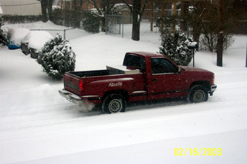Red regular-cab Chevrolet pickup parked on snowy street during snowfall