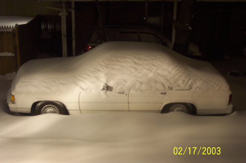 Sedan completely covered in snow parked outside at night near a fence