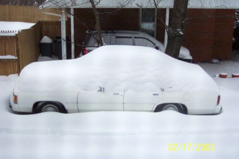 1991 Ford Crown Victoria LTD - A white sedan is almost completely covered by a thick layer of snow, with another vehicle partially visible behind it.