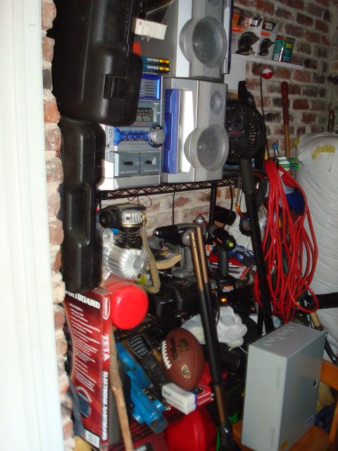 1977 Ford F-100 - A disorganized storage area filled with various items, including electronics, tools, and sports equipment, is visible against a brick wall.
