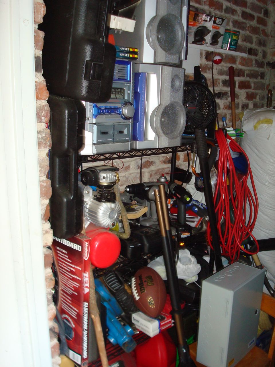 Cluttered storage area with shelves holding audio equipment, tools, an air compressor, and sports gear against a brick wall