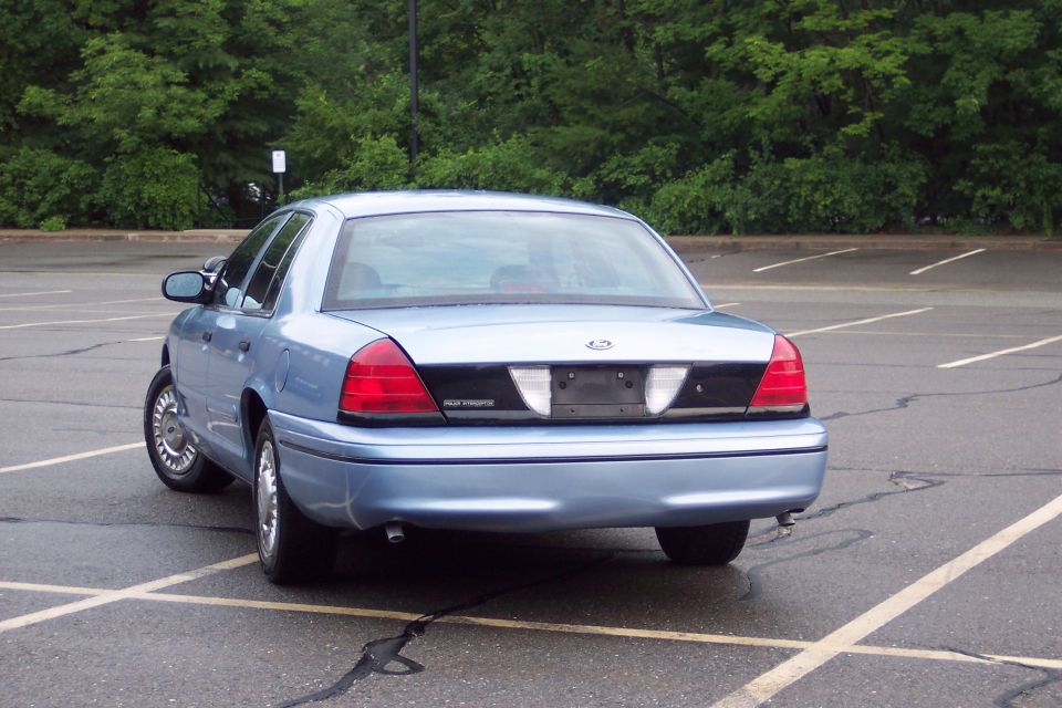 Light blue 2001 Ford Crown Victoria sedan, rear passenger side view, parked in a lot.