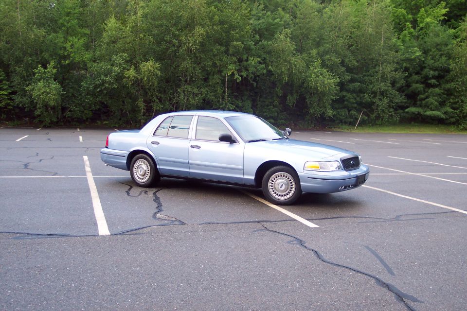 Light blue 2001 Ford Crown Victoria sedan parked in an asphalt lot with trees in the background.
