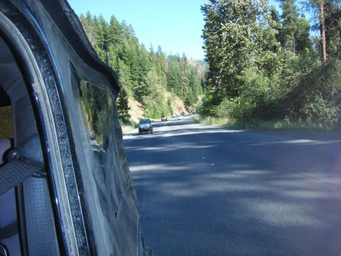 2005 Jeep Wrangler - A dark colored Jeep Wrangler is driving on a paved road through a forest with tall evergreen trees. The photo is taken from inside another…