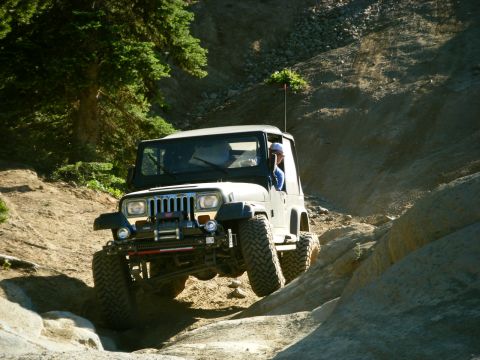 2005 Jeep Wrangler - A dark-colored Jeep Wrangler is navigating a rocky, off-road trail with a driver visible inside. The Jeep is angled upwards, with its front…