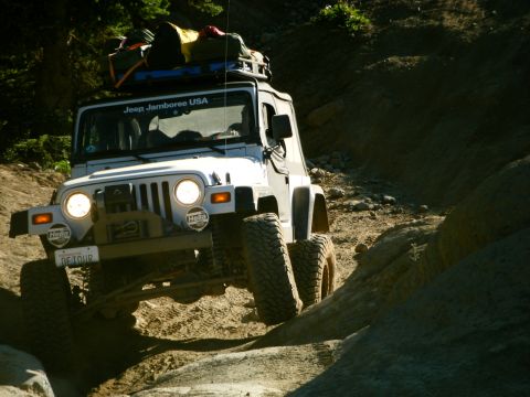 2005 Jeep Wrangler - A white Jeep Wrangler is driving up a dirt trail, navigating over rocks and uneven terrain. The vehicle has a roof rack loaded with gear an…