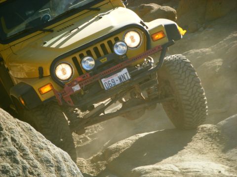 2005 Jeep Wrangler - A yellow Jeep Wrangler is shown from a low angle, navigating a rocky, off-road terrain. The vehicle has a red Hi-Lift jack mounted on the f…
