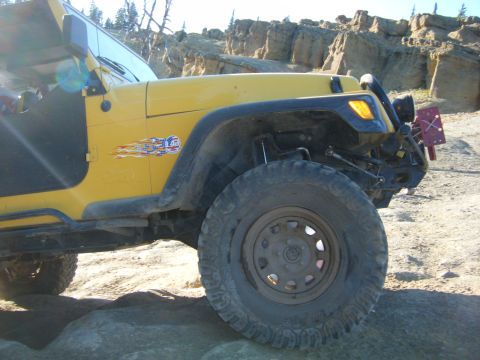 2005 Jeep Wrangler - A yellow Jeep Wrangler is parked on a rocky, dirt trail with a cliff face in the background. The vehicle has an American flag decal with fl…