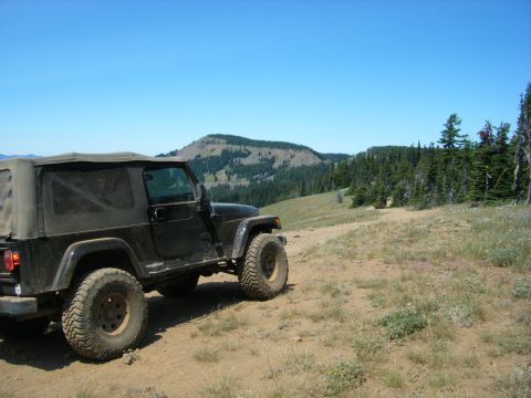 2005 Jeep Wrangler - A black Jeep Wrangler is parked on a dirt road with a wooded mountain landscape in the background under a clear blue sky.