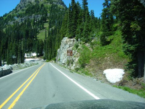 2005 Jeep Wrangler - A winding mountain road leads past a sign for Tipsoo Lake and a snow patch, with a steep, forested mountainside on the right.