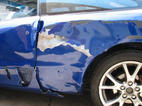 1993 Ford Probe - The side of a blue car shows significant damage to the door and quarter panel, with peeling paint revealing rust and metal underneath. The…